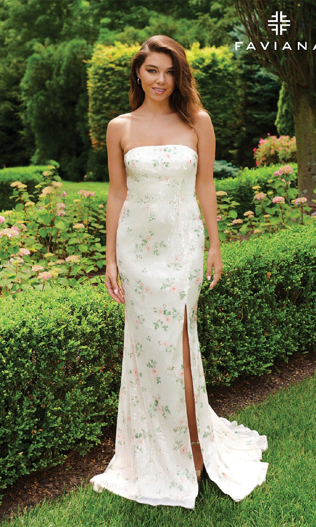 A woman stands in a garden wearing the Faviana 11222 Floral-Print Sequin Long Prom Dress featuring a strapless design and thigh-high slit.